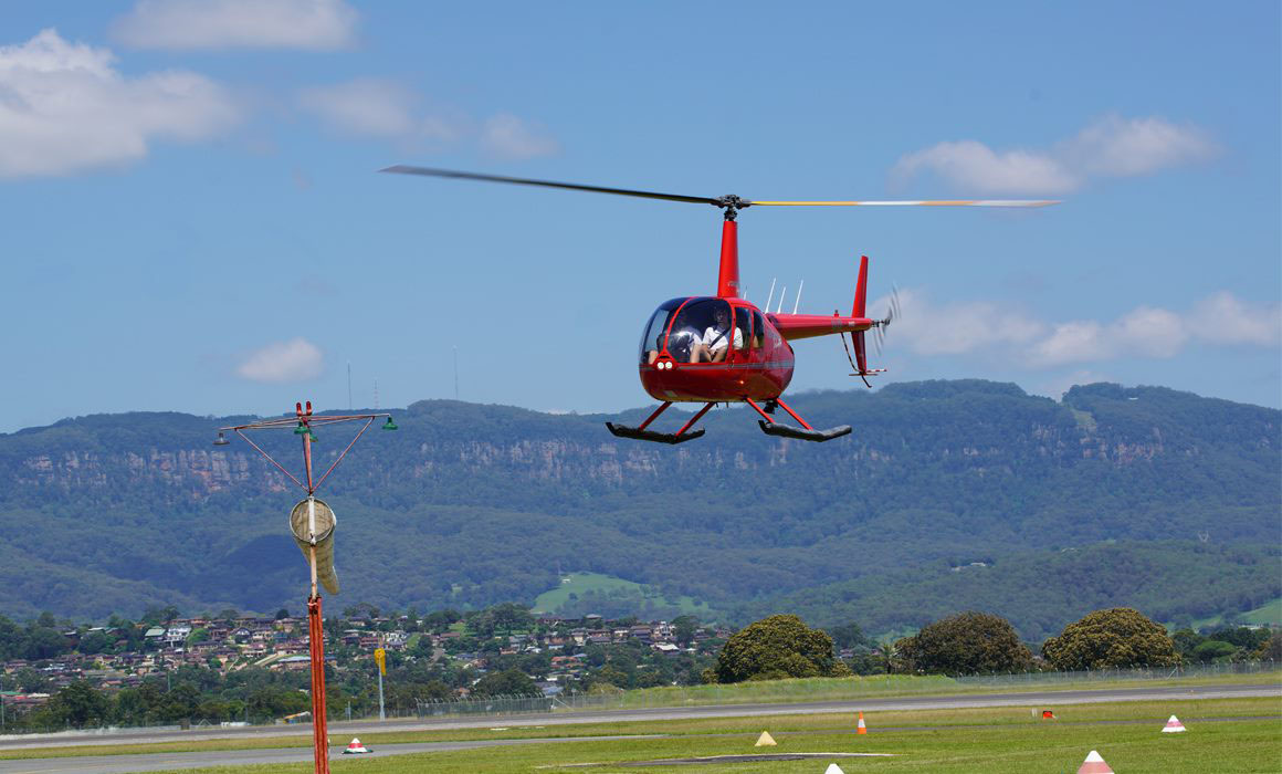 A red Robinson R44 helicopter landing at Shellharbour Airport near to a windsock