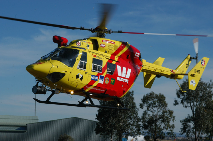 A yellow-and-red helicopter with the Westpac bank logo printed on it flying low to the ground, with a building and some trees visible in the background