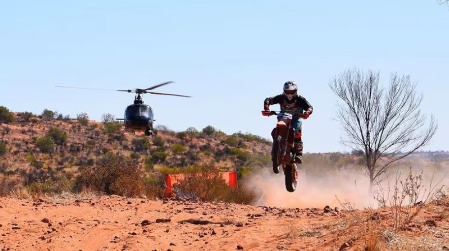 A helicopter sits in the background as a motorcyclist drives along a dirt road in front of it