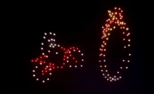 A swarm of drones in the night sky, making the image of a man on a motorbike driving through a ring of fire with their lights