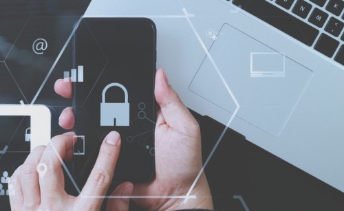 Hands holding a mobile phone with a lock symbol over it, with a laptop in the background