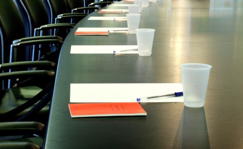 A long desk prepared for a meeting with paper, pens, and cups placed in front of office chairs