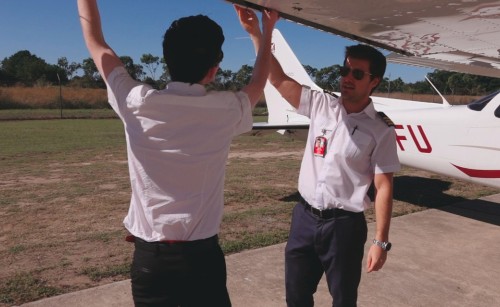 Two pilots under the wing of a small aircraft, reaching up to touch it and facing each other.