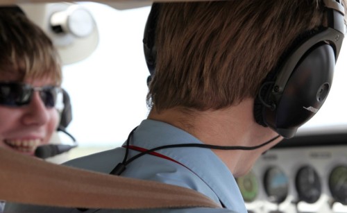 The back of the head of a flight instructor, with a student pilot sitting next to them in the cockpit of a plane