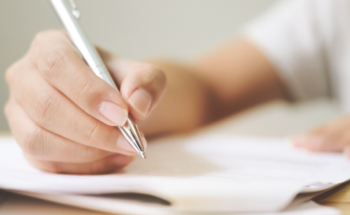 A hand holding a metalling pen, holding it over a stack of folded-over papers.