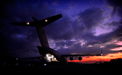 An aeroplane with its rear cargo door open and interior lights on as the sun sets behind it