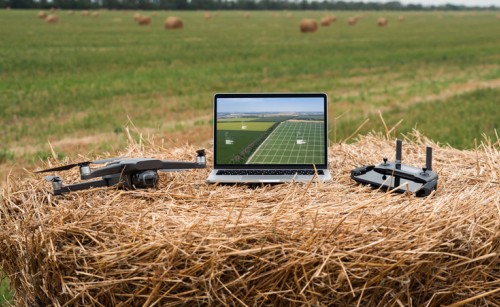 A laptop sitting on a bale of hay in a field, with a drone and a drone controller sitting on either side of it