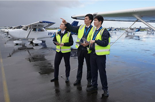 Three pilots wearing high-visibility vests, standing on a wet taxiway with a plane behind them