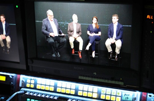 A screen with a keyboard underneath it, displaying four aviation experts sitting on chairs in a dark room