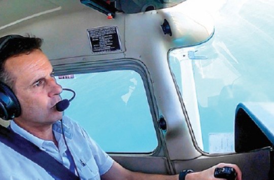 A pilot flying a plane, with blue sky visible through the windows behind him