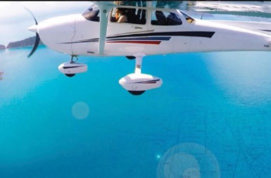 A white plane with stripes on its side is flying above bright blue water with a few islands in the background.