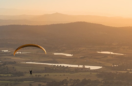 A man parachuting over fields and lakes, with mist-covered mountains in the background