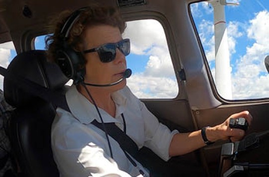 A female pilot flying a plane, with fluffy blue clouds visible through the windows behind her