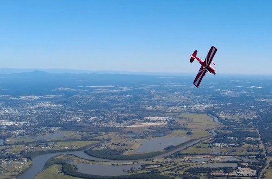 An aerial shot of a red plane doing aerobatics over a green landscape dotted with buildings, and the blue sky behind it
