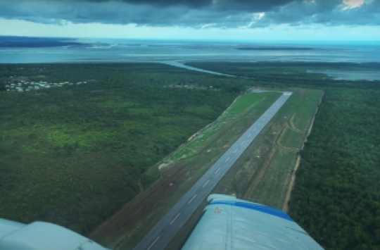 A view of an airstrip over the edge of an airplane's wing, surrounded by green bushland and with the ocean in the distance behind it