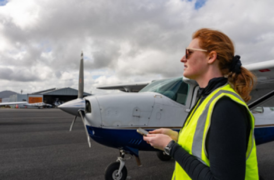 A woman wearing a high visibility vest looks out across an airport with an aeroplane parked behind her