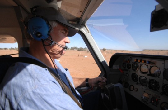Man wearing headset flying a small aircraft over dry outback terrain.