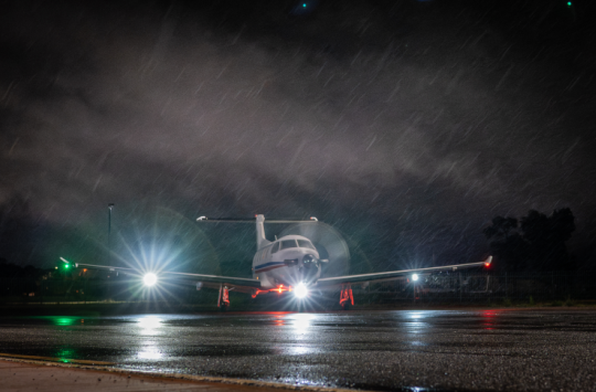 Light aircraft with headlights on sits on a wet runway at night in rain.