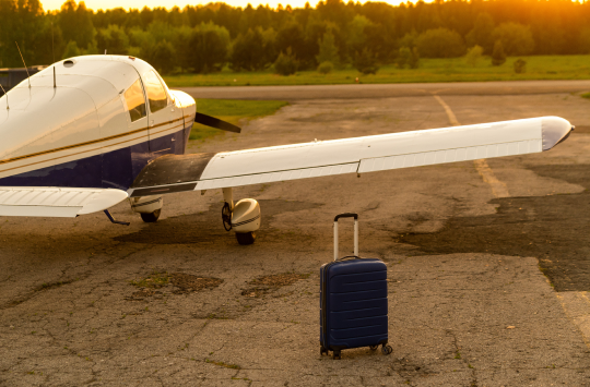 A blue suitcase and a parked small private jet. Quadruple plane with a propeller for an air taxi in the sunset. 