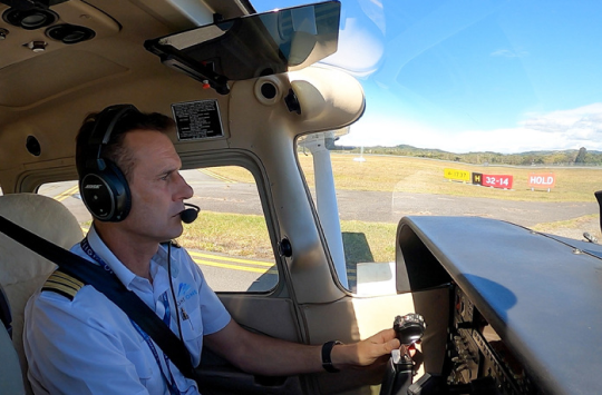 Pilot in a light aircraft cockpit at a controlled aerodrome, wearing a headset and preparing for taxi, with the runway visible ahead.