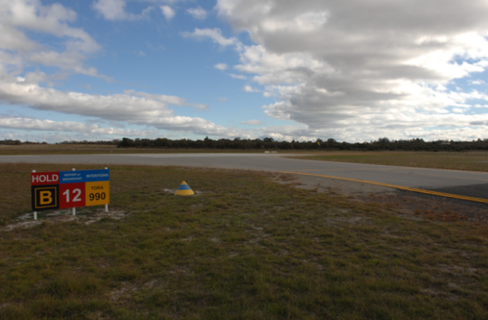 View of taxiway B and runway 12 at Jandakot Aerodrome, showing a runway holding point sign and the intersection area where pilots must maintain situational awareness while taxiing.
