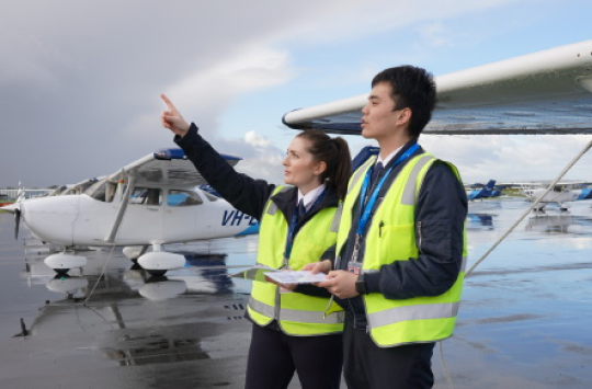 Two pilots in high-visibility vests stand beside a light aircraft on a wet tarmac, looking towards approaching dark clouds.