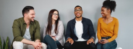 A white man and woman sitting next to a black man and woman in a row of chairs; the black man looks upward hopefully while the others look at him and smile