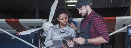 A white man and black woman standing in a hangar examining tablets, with an aeroplane being maintained behind them