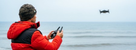 A ,am om a red jumper and a beanie hat flies a drone on the beach, with the ocean in the background