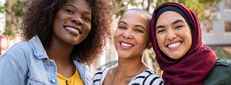 Three woman stand in a row, smiling. The one on the right wears a Muslim headdress while the one in the middle is bald.