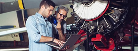 Maintenance engineers looking up documentation on a laptop with an aircraft engine behind them