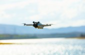 A white RPA flying with a lake in the background