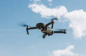 A drone flying through the blue sky, with a cloud behind it