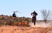 A helicopter sits in the background as a motorcyclist drives along a dirt road in front of it