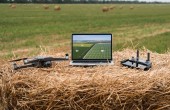 A laptop sitting on a bale of hay in a field, with a drone and a drone controller sitting on either side of it