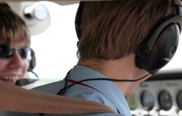 The back of the head of a flight instructor, with a student pilot sitting next to them in the cockpit of a plane
