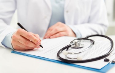 The body and hands of a doctor in a white coat writing on paper held by a clipboard, with a stethoscope sitting on top of the page.