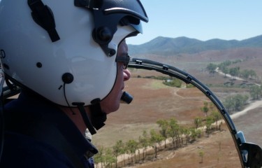 A pilot with a white helmet flies a helicopter; the Outback can be seen through his cockpit window