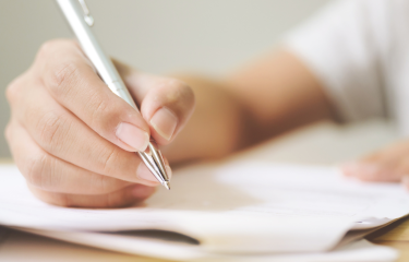 A hand holding a metalling pen, holding it over a stack of folded-over papers.