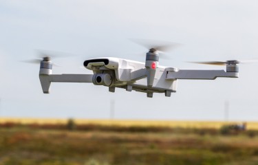 A white drone flying through the air, with a paddock in the background