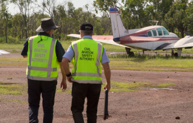 Two CASA inspectors wearing high-visibility vests with "Civil Aviation Safety Authority" written on them examining a plane parked on grass beside a dirt track