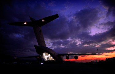 An aeroplane with its rear cargo door open and interior lights on as the sun sets behind it