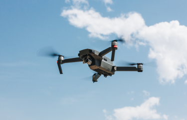 A drone flying through the blue sky, with a cloud behind it